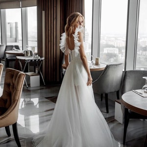 K&ouml;nnte beinhalten: Wei&szlig;es Hochzeitskleid mit T&uuml;llrock und R&uuml;schenausschnitt. Das Kleid hat einen tiefen R&uuml;cken und ein eng anliegendes Oberteil. Die Braut steht in einem Restaurant mit gro&szlig;en Fenstern mit Blick auf eine Stadt.