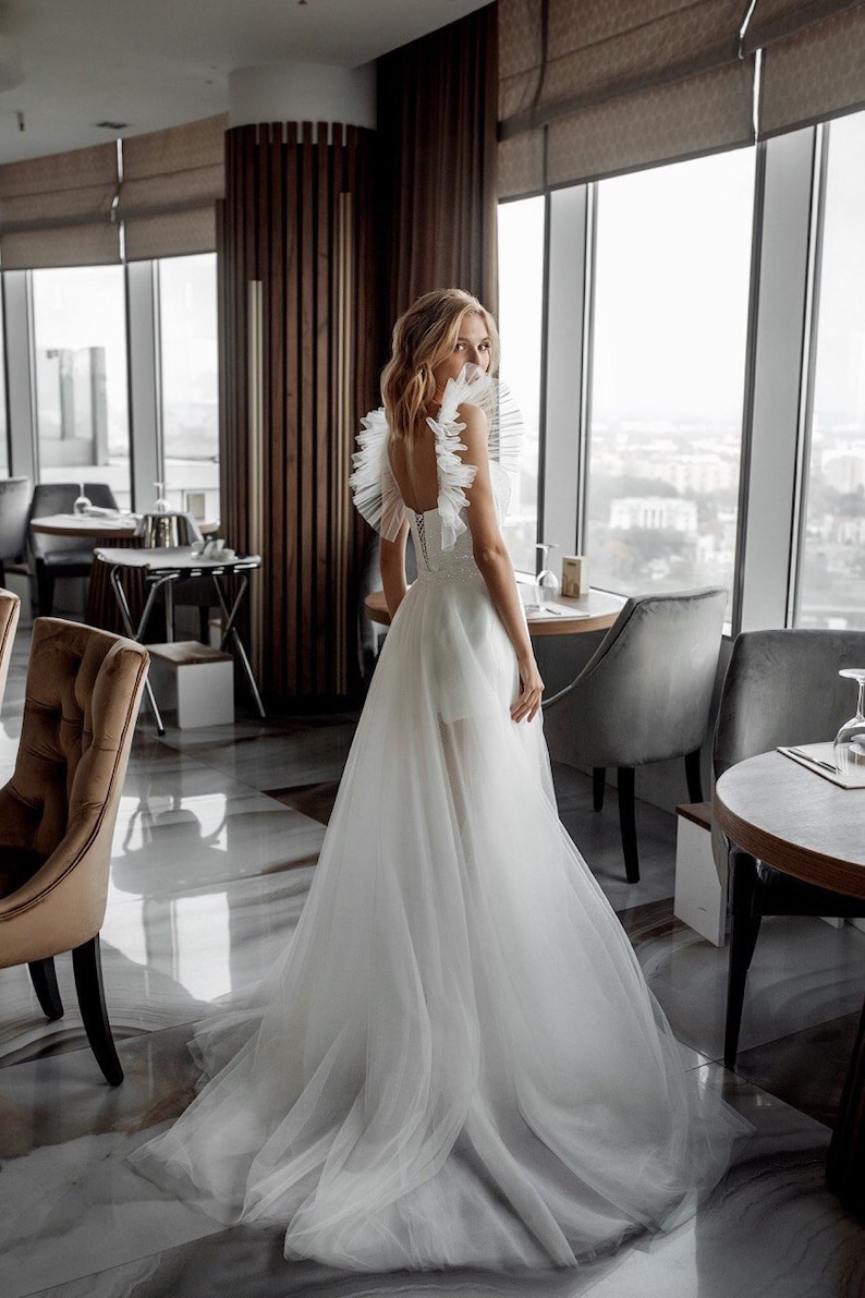 K&ouml;nnte beinhalten: Wei&szlig;es Hochzeitskleid mit T&uuml;llrock und R&uuml;schenausschnitt. Das Kleid hat einen tiefen R&uuml;cken und ein eng anliegendes Oberteil. Die Braut steht in einem Restaurant mit gro&szlig;en Fenstern mit Blick auf eine Stadt.