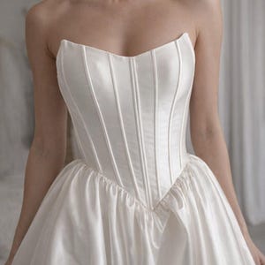 May include: A strapless white satin wedding dress with a structured bodice and a full skirt. The bodice features vertical seams for a tailored fit. The dress is elegant and classic, suitable for a bridal occasion.