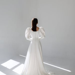 May include: A white wedding dress with a fitted bodice and a flowing skirt. The dress has a square neckline and long, puffed sleeves. The back of the dress features a row of buttons.