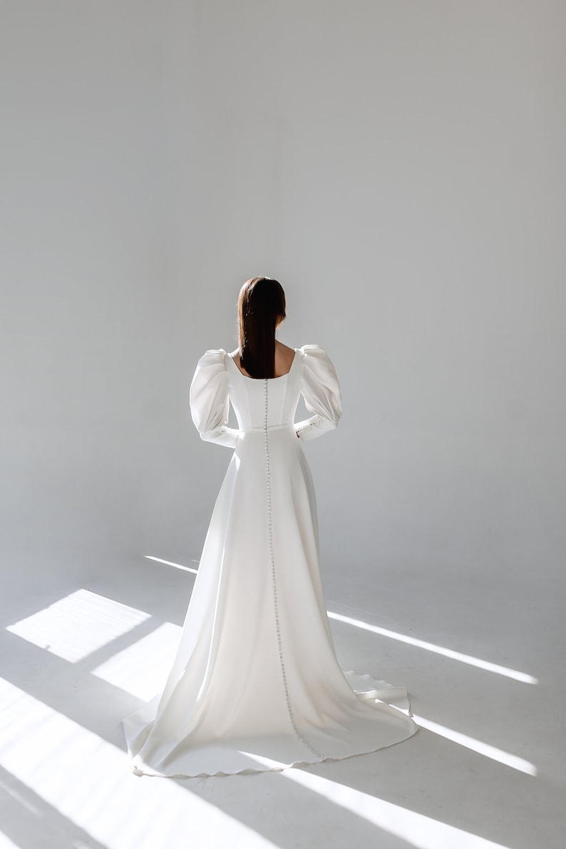May include: A white wedding dress with a fitted bodice and a flowing skirt. The dress has a square neckline and long, puffed sleeves. The back of the dress features a row of buttons.