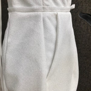 K&ouml;nnte beinhalten: Nahaufnahme eines wei&szlig;en, glitzernden Jumpsuits. Der Stoff hat ein strukturiertes Aussehen und einen subtilen Schimmer. Der Jumpsuit hat eine betonte Taille mit passendem G&uuml;rtel und ein drapiertes Design am Oberteil.