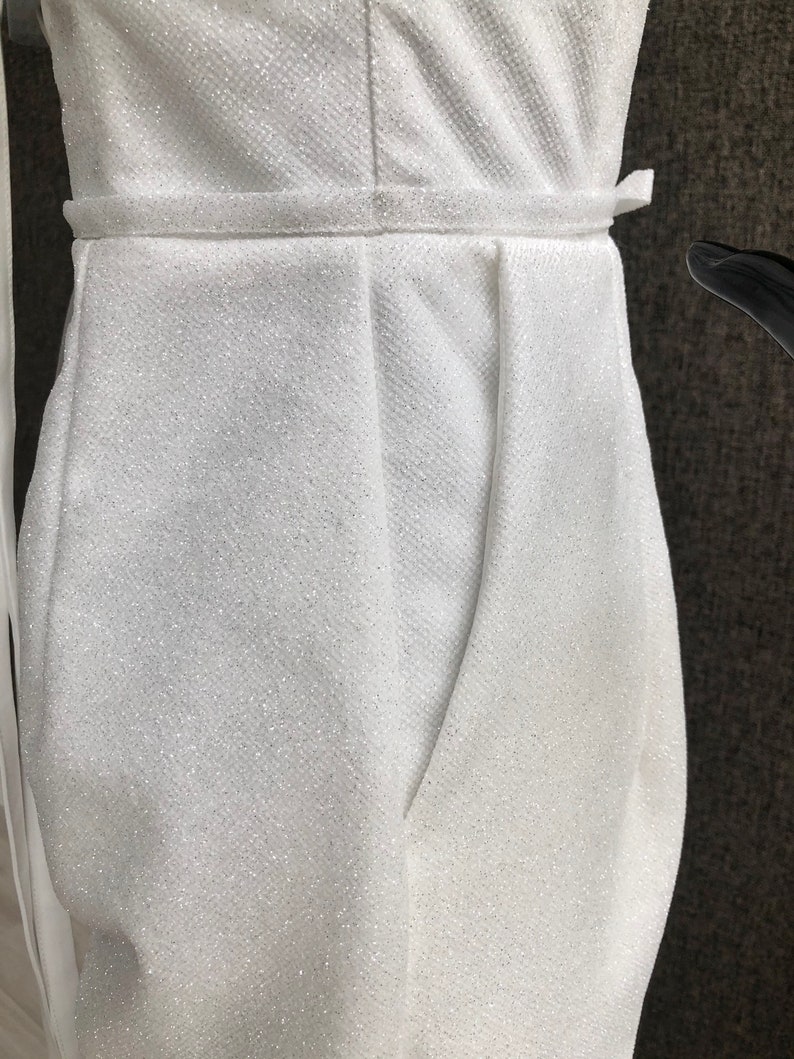 K&ouml;nnte beinhalten: Nahaufnahme eines wei&szlig;en, glitzernden Jumpsuits. Der Stoff hat ein strukturiertes Aussehen und einen subtilen Schimmer. Der Jumpsuit hat eine betonte Taille mit passendem G&uuml;rtel und ein drapiertes Design am Oberteil.