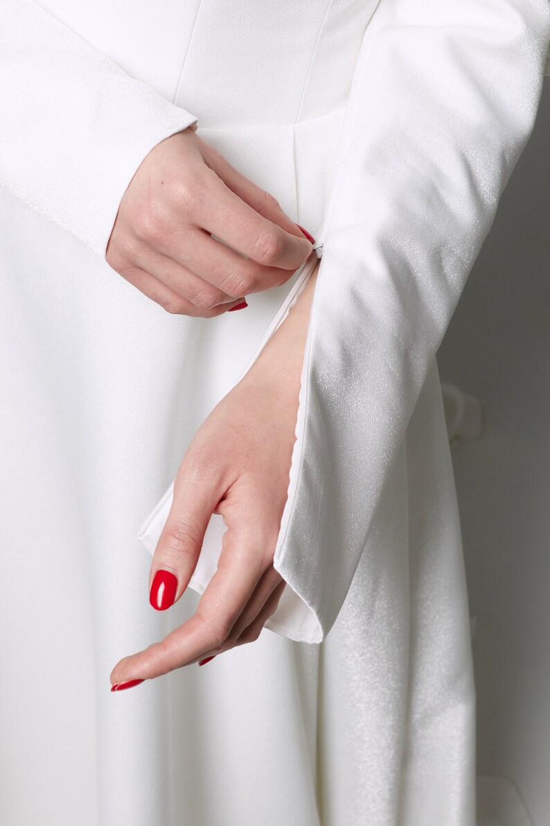 Puede incluir: Un vestido blanco con mangas largas y un detalle de cremallera en el pu&ntilde;o. Las mangas est&aacute;n fruncidas en la mu&ntilde;eca y el vestido tiene una falda fluida.