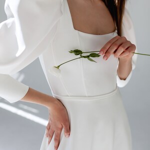 May include: A white wedding dress with a square neckline and puffy sleeves. The dress has a fitted bodice and a flowing skirt. The model is holding a single white flower.