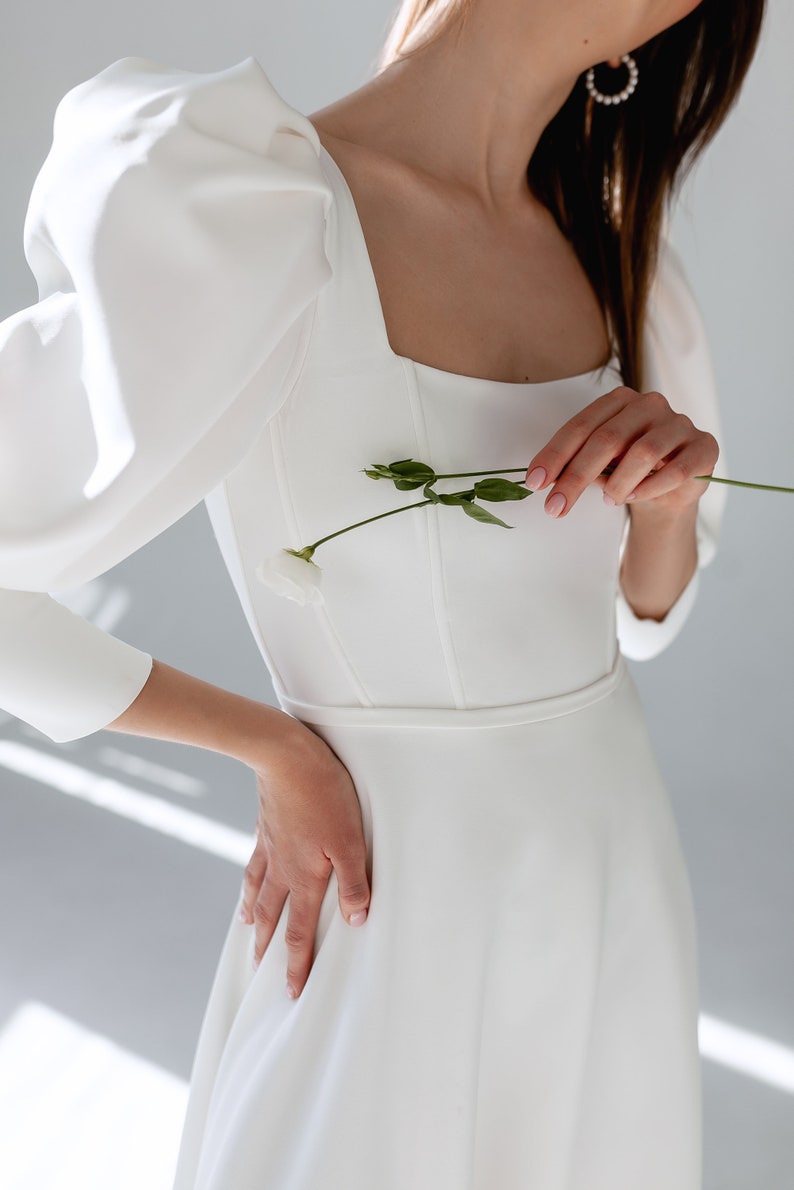 May include: A white wedding dress with a square neckline and puffy sleeves. The dress has a fitted bodice and a flowing skirt. The model is holding a single white flower.