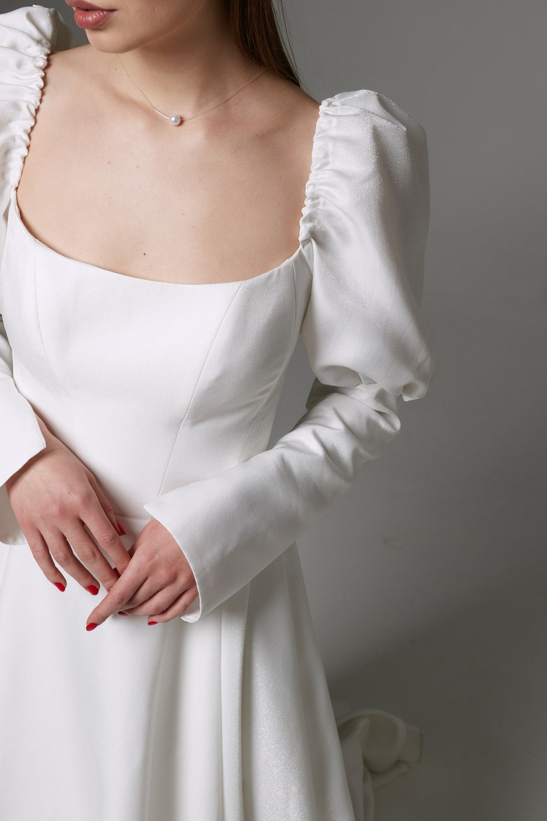 Puede incluir: Un vestido de novia blanco con escote cuadrado y mangas largas. Las mangas son abullonadas en los hombros y tienen un pu&ntilde;o ajustado. El vestido tiene un corpi&ntilde;o ajustado y una falda fluida.
