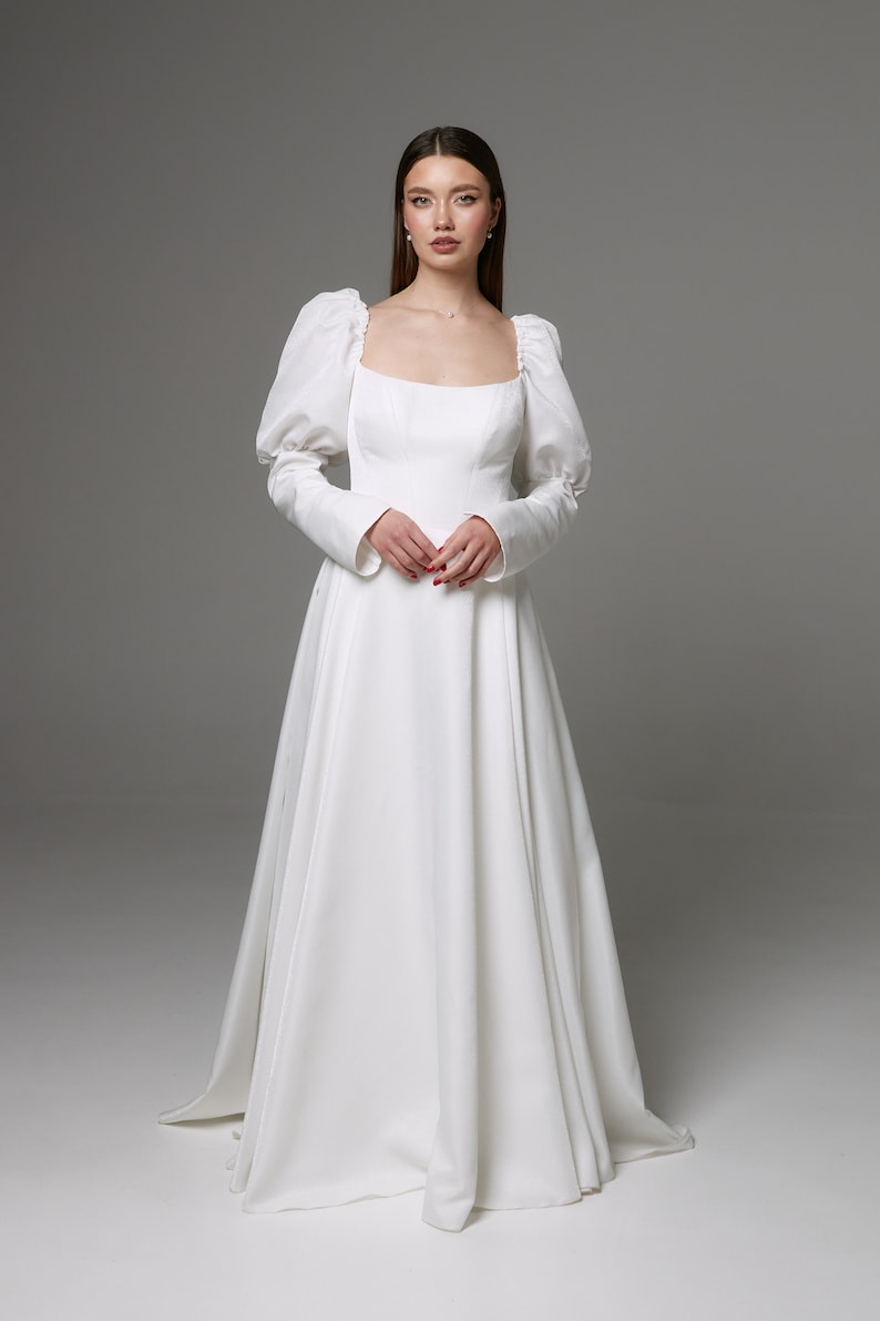 Puede incluir: Un vestido de novia blanco con escote cuadrado y mangas largas abullonadas. El vestido tiene un corpi&ntilde;o ajustado y una falda fluida.