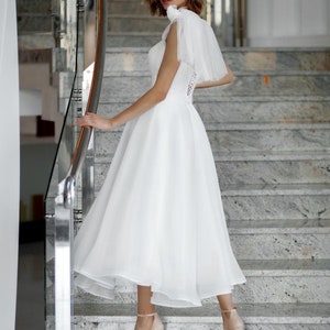 May include: A white wedding dress with a tulle skirt and a bodice with lace detailing. The dress is paired with nude-colored platform heels. The model is standing on a marble staircase, posing for a photo.