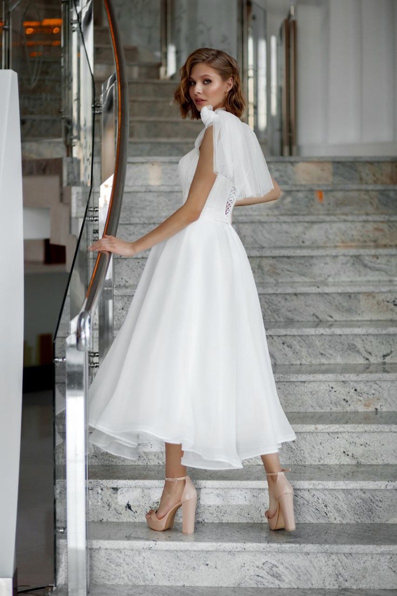 May include: A white wedding dress with a tulle skirt and a bodice with lace detailing. The dress is paired with nude-colored platform heels. The model is standing on a marble staircase, posing for a photo.