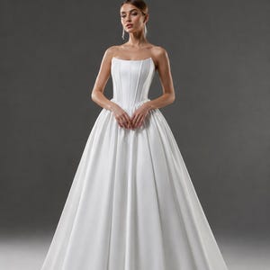 Könnte beinhalten: Ein trägerloses weißes Satin-Brautkleid mit einem eng anliegenden Oberteil und einem vollen, fließenden Rock. Das Kleid hat vertikale Nähte am Oberteil und eine dezente Schleppe. Das Model trägt baumelnde Ohrringe.