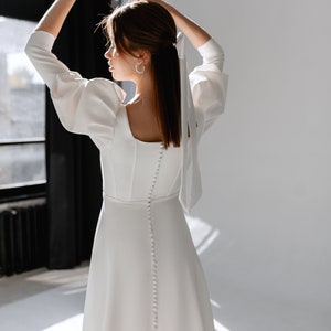 May include: A white dress with long sleeves and a button-up back. The dress has a square neckline and a fitted bodice. The model is wearing the dress with her hair pulled back in a ponytail.