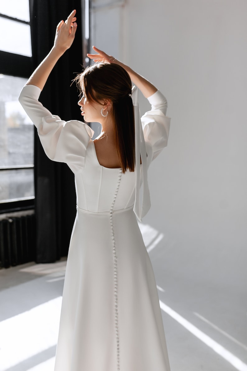 May include: A white dress with long sleeves and a button-up back. The dress has a square neckline and a fitted bodice. The model is wearing the dress with her hair pulled back in a ponytail.