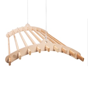 May include: A natural wood, hanging drying rack with a curved design. The rack features multiple dowels for hanging clothes and is suspended by a white rope. The rack is made of light-colored wood and has a minimalist aesthetic.