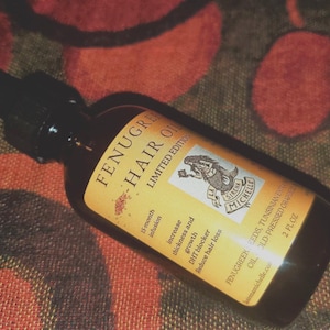 May include: A glass bottle of Fenugreek Hair Oil with a yellow label. The label features a mermaid illustration and text that reads "Fenugreek Seeds, Tunisian Oil Cold Pressed Craft, 15 month infusion, Increase thickness and growth, DHT blocker, Reduce hair loss, 2 fl oz, Sirena Michelle, mamichelle.co".