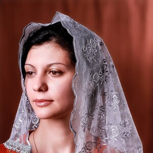 May include: A woman wearing a silver lace veil with floral embroidery. The veil is draped over her head and shoulders, and she is wearing a red dress with a lace collar.
