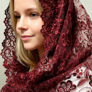 May include: A burgundy lace head covering with floral embroidery. The intricate design features delicate flowers and swirling patterns. The head covering is draped over a person's head and shoulders, showcasing the detailed craftsmanship.