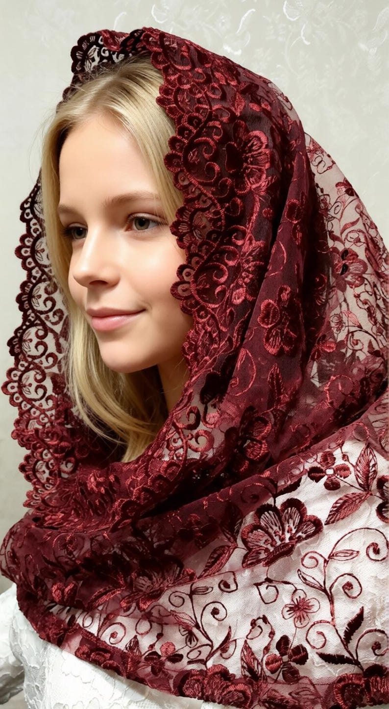 May include: A burgundy lace head covering with floral embroidery. The intricate design features delicate flowers and swirling patterns. The head covering is draped over a person's head and shoulders, showcasing the detailed craftsmanship.