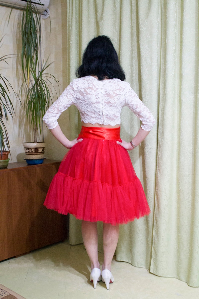 May include: A woman wearing a white lace crop top with a red satin ribbon and a red tulle skirt. She is standing in front of a white curtain.