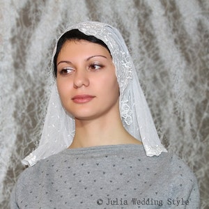 May include: A white lace veil with a floral pattern. The veil is worn by a person with dark hair and is covering their head and shoulders.