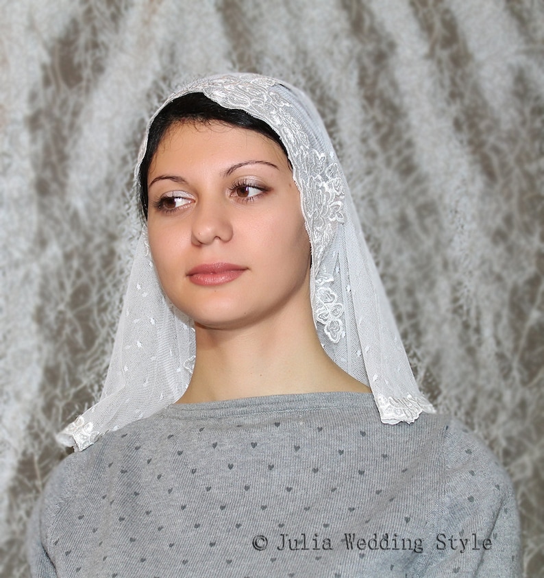 May include: A white lace veil with a floral pattern. The veil is worn by a person with dark hair and is covering their head and shoulders.