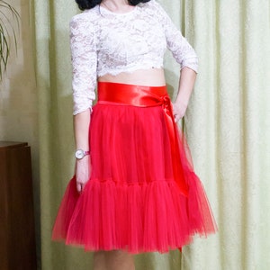 May include: A woman wearing a white lace top with a red satin belt and a red tulle skirt. She is wearing white heels.