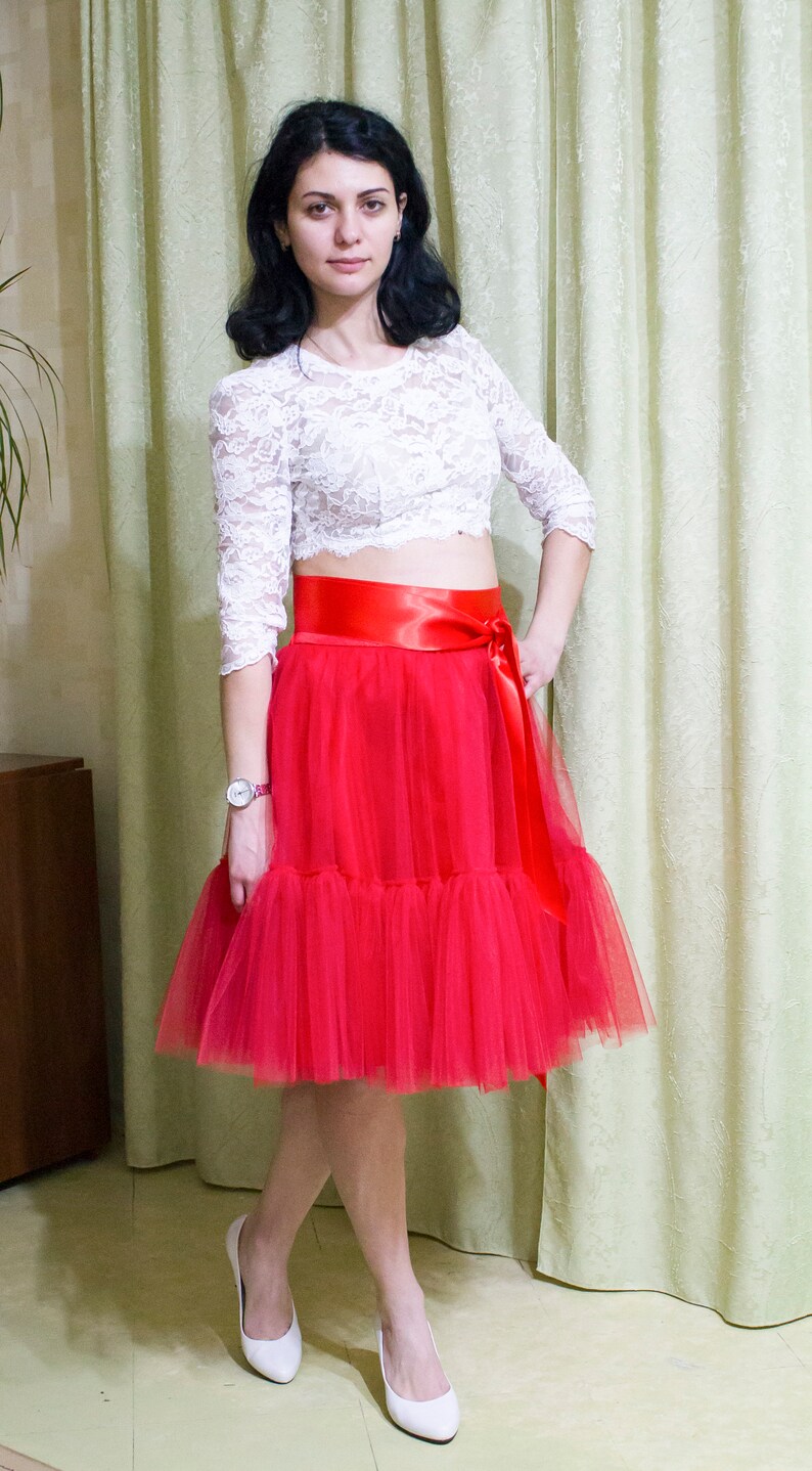 May include: A woman wearing a white lace top with a red satin belt and a red tulle skirt. She is wearing white heels.