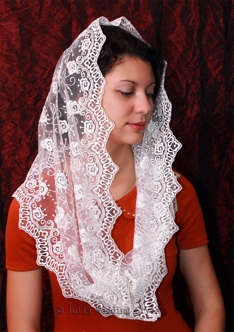 May include: White lace mantilla with floral embroidery. The lace is delicate and has a scalloped edge. The mantilla is worn over a red dress.
