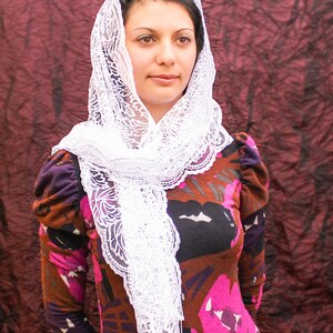 May include: A woman wearing a white lace veil over a brown and pink floral print dress. The veil is draped over her head and shoulders, and she is looking at the camera.