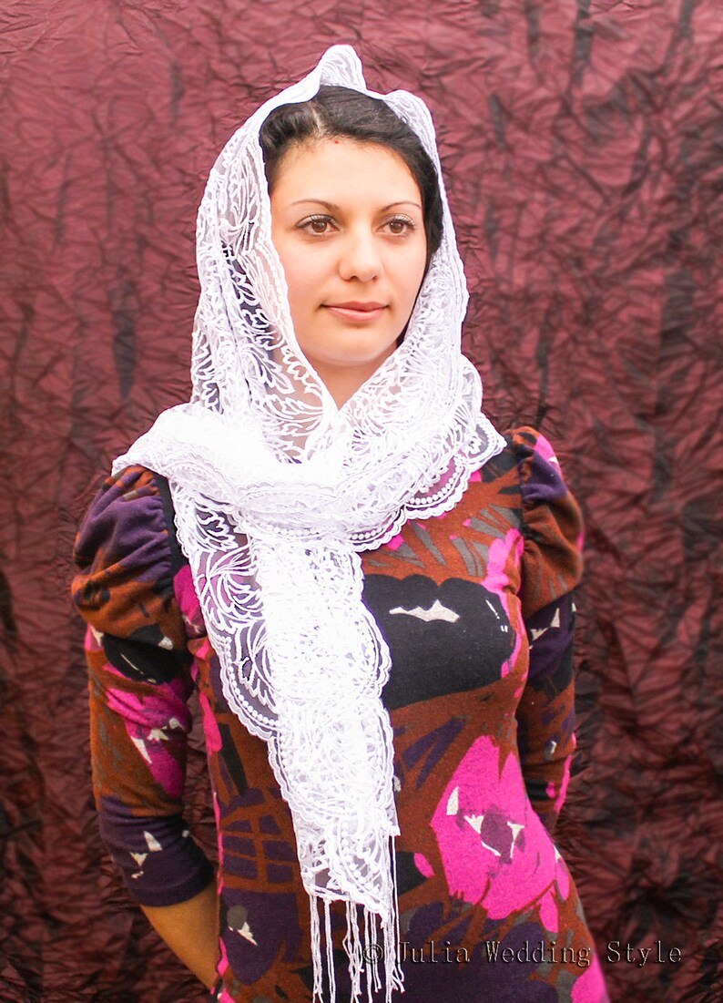 May include: A woman wearing a white lace veil over a brown and pink floral print dress. The veil is draped over her head and shoulders, and she is looking at the camera.