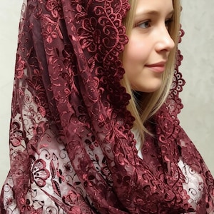 May include: A burgundy lace head covering with floral patterns. The delicate fabric drapes over the head and shoulders, creating a soft, elegant silhouette. The intricate design and rich color suggest a formal or religious context.