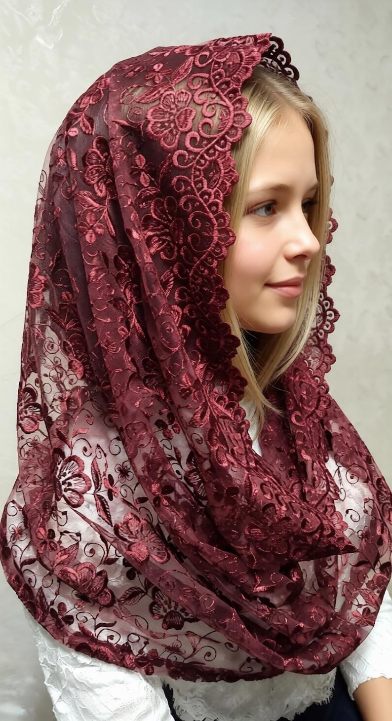 May include: A burgundy lace head covering with floral patterns. The delicate fabric drapes over the head and shoulders, creating a soft, elegant silhouette. The intricate design and rich color suggest a formal or religious context.
