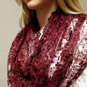 May include: A burgundy lace infinity scarf with a floral pattern. The scarf is draped around the neck, showcasing the intricate details of the lace. The scarf is made of a soft, sheer material.