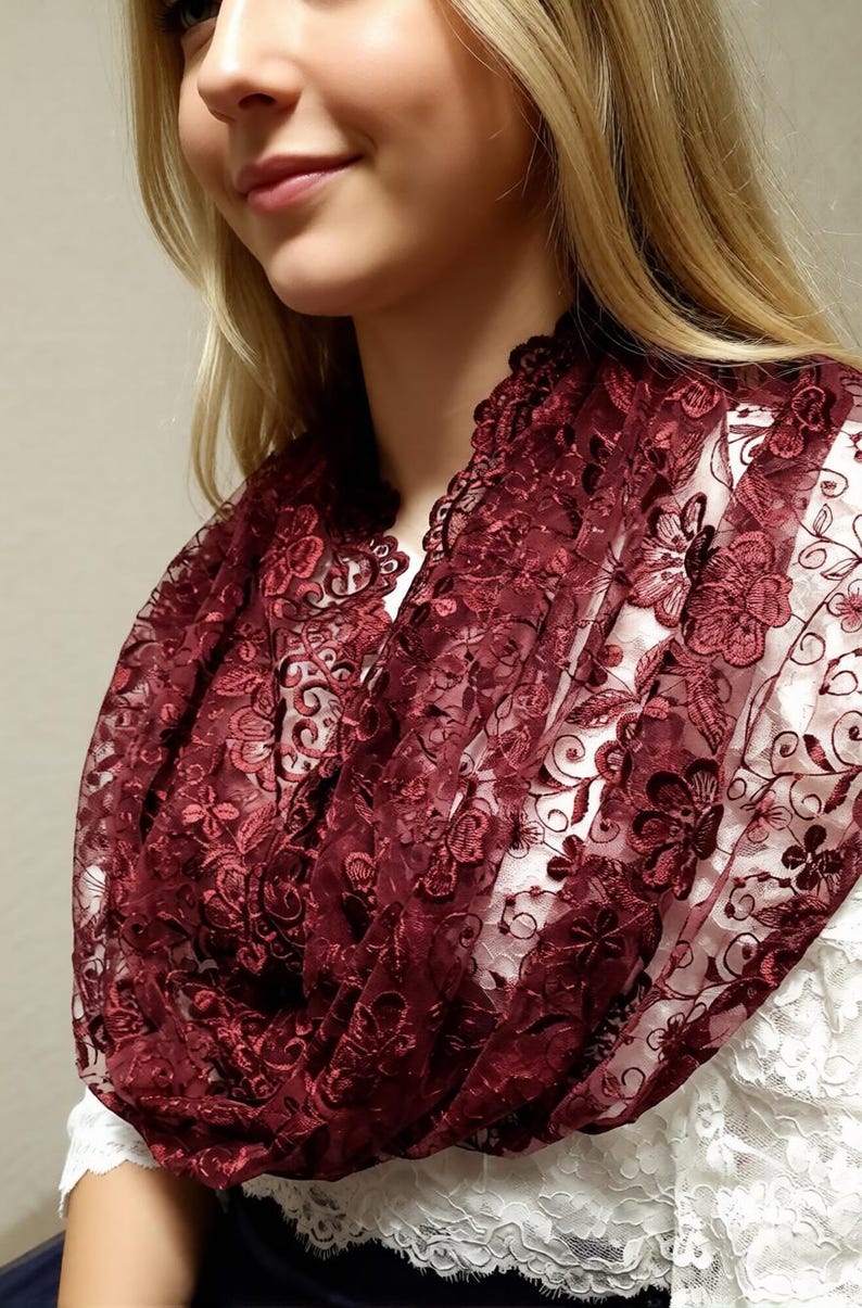 May include: A burgundy lace infinity scarf with a floral pattern. The scarf is draped around the neck, showcasing the intricate details of the lace. The scarf is made of a soft, sheer material.