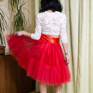 May include: A woman wearing a white lace top with a red tulle skirt and a satin ribbon waist. She is wearing white heels.