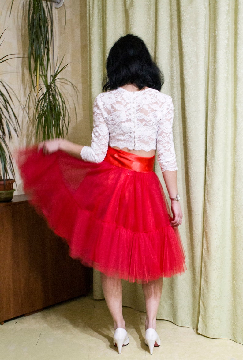 May include: A woman wearing a white lace top with a red tulle skirt and a satin ribbon waist. She is wearing white heels.