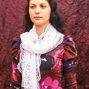 May include: A woman with long dark hair wears a white lace scarf over a floral print dress with a dark background. The dress has a pink floral pattern with brown and purple accents. The woman is looking to the side.