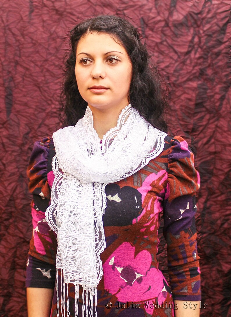 May include: A woman with long dark hair wears a white lace scarf over a floral print dress with a dark background. The dress has a pink floral pattern with brown and purple accents. The woman is looking to the side.