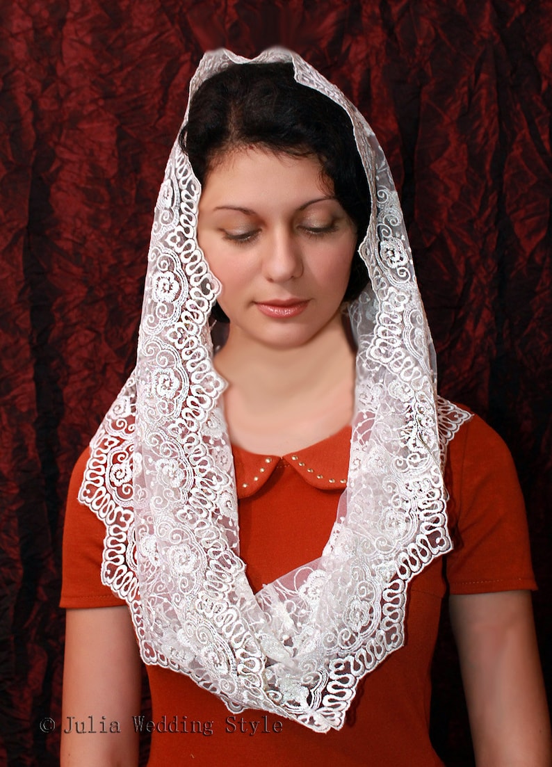May include: A white lace veil with a scalloped edge and floral embroidery. The veil is worn by a person with dark hair and is draped over their shoulders.