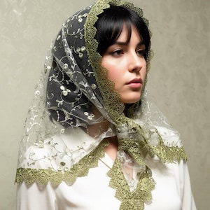 Puede incluir: Un velo transparente bordado en verde oliva y blanco con un ribete de encaje festoneado. El velo está drapeado sobre una camisa blanca con cuello. El bordado presenta patrones florales y de hojas.