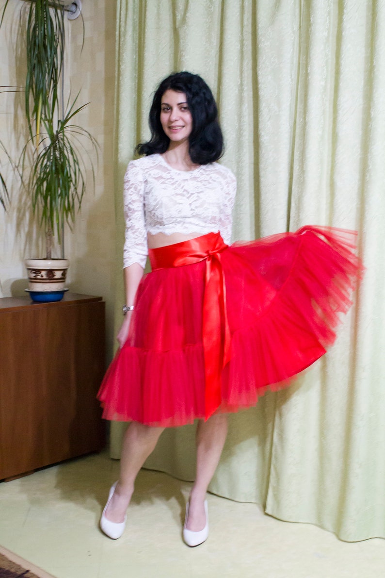 May include: A woman wearing a white lace crop top with a red satin ribbon tied around the waist, a red tulle skirt, and white heels.