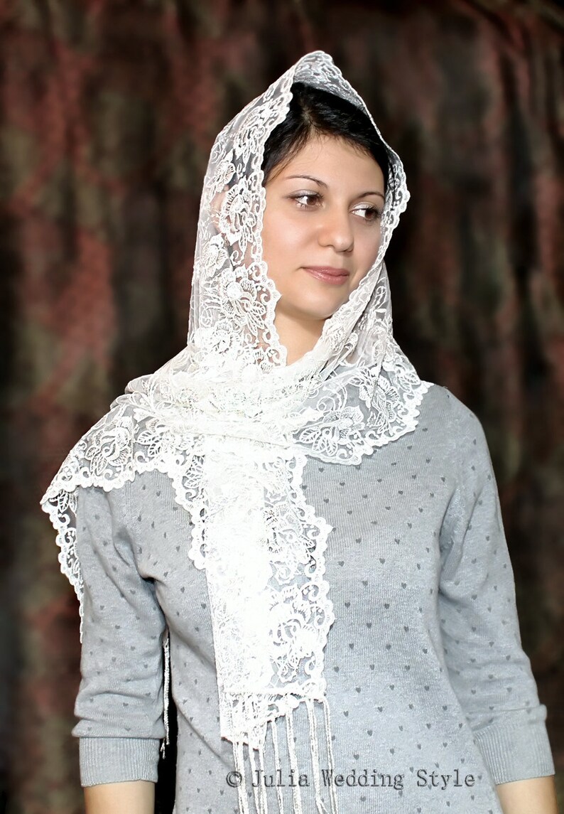 Pu&ograve; includere: Una donna indossa un velo di pizzo bianco su un maglione grigio con un motivo a forma di cuore. Il velo &egrave; drappeggiato sulla testa e sulle spalle, e il pizzo &egrave; delicato e intricato.