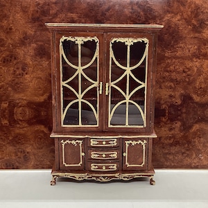 May include: Miniature wooden cabinet with glass doors and gold accents. The cabinet has two doors on top and three drawers below. The dark brown wood is complemented by the gold trim and decorative details, creating a classic look.