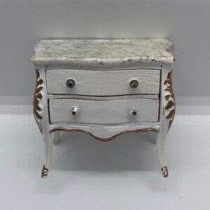 May include: White painted wooden chest of drawers with gold accents and marble top. The chest has two drawers with silver knobs.