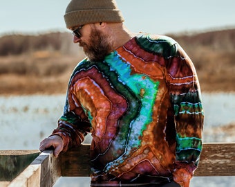 May include: A person wearing a brown beanie and a long-sleeved shirt with a colorful tie-dye pattern. The shirt has a swirling design of orange, red, green, blue, and purple. The person is standing on a wooden railing overlooking a body of water.