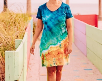 May include: A short-sleeved V-neck dress featuring a tie-dye pattern that transitions from deep blue at the top to a sandy tan color at the bottom, separated by a white line that mimics the appearance of a shoreline meeting the ocean water.