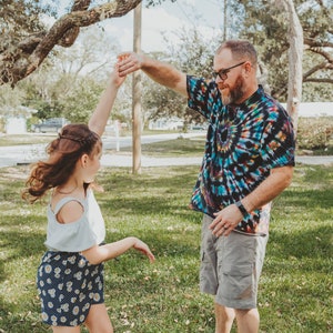 May include: A father and daughter are dancing in a grassy park. The father is wearing a tie-dye shirt and khaki shorts. The daughter is wearing a white top and a floral skirt. They are both smiling and having fun.