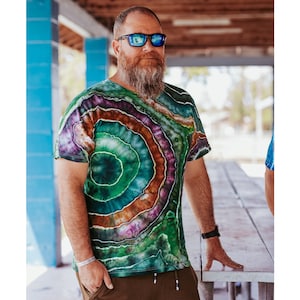 Bearded man with reflective sunglasses standing under a covered porch. he is wearing a geode tie dye shirt. It is a short sleeve cotton, ice dyed in greens, brown and purple.