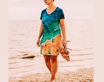 May include: A short-sleeved V-neck dress featuring a tie-dye pattern that transitions from deep blue and teal at the top to a sandy orange and tan color at the bottom, mimicking a coastal landscape with a clear line separating the colors.