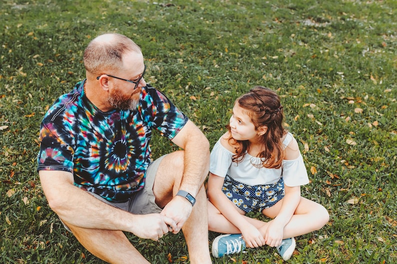 May include: A man and a girl are sitting on the grass, looking at each other. The man is wearing a tie-dye shirt and the girl is wearing a white shirt with a floral pattern.